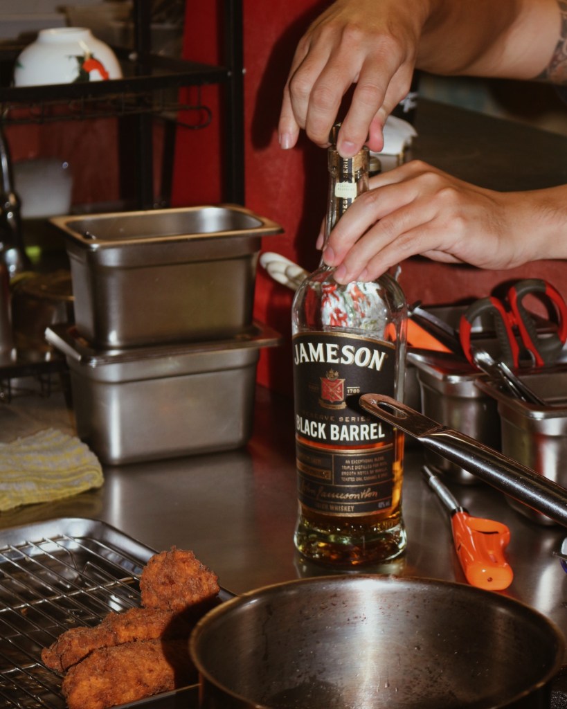 A close-up of hands opening a bottle of Jameson Black Barrel whiskey in a kitchen setting, with fried food in the foreground and various kitchen utensils in the background.