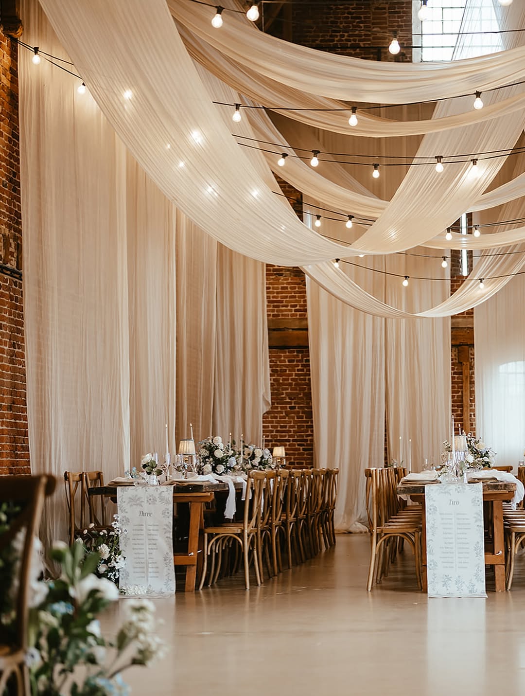 Elegant wedding reception decor with draped fabric, string lights, long wooden tables, and flower centerpieces.