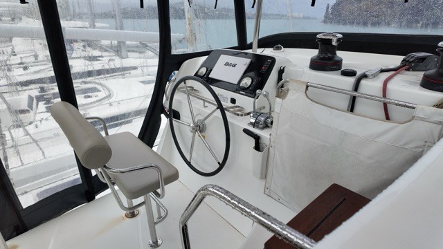 Lagoon 42 catamaran helm station showing the stainless steel steering wheel, B&G chartplotter and instrument display, self-tailing sheet winch, helm seat on a folding stainless bracket, teak helm pod step, and panoramic black-framed windscreen glazing looking out over a snow-dusted winter marina