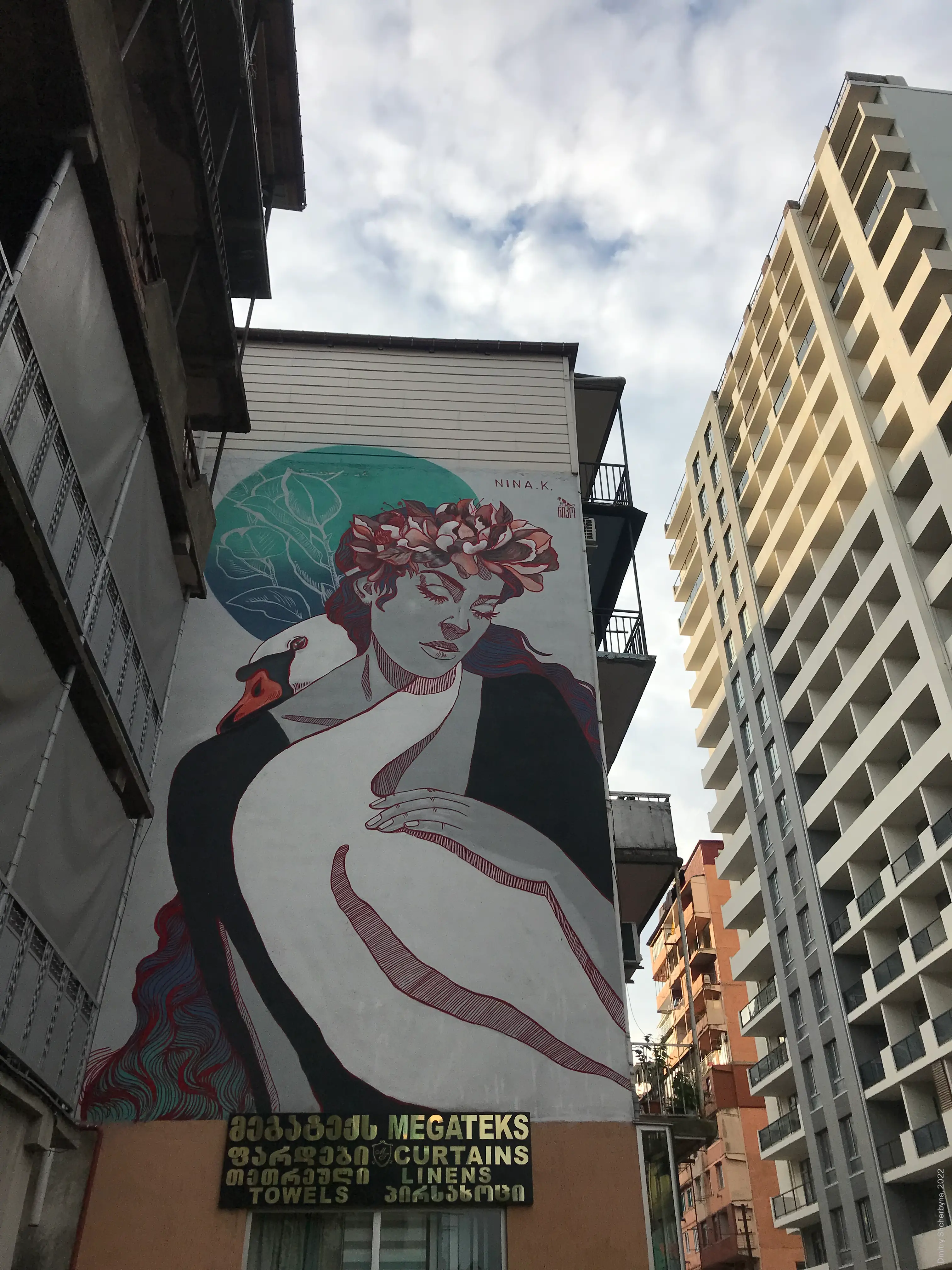 A mural of a woman with closed eyes and flowers in her dark red hair, gently embracing a white swan against a teal circular background. The style is inspired by art nouveau with fine red linework on pale skin.