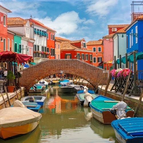 Canal with small boats, colorful buildings, and a brick bridge in a bright, clear-weathered town. Flowers on the right side.