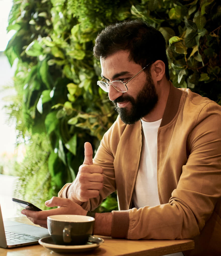 A bearded man with glasses and a laptop sitting at a table, giving a thumbs up gesture.