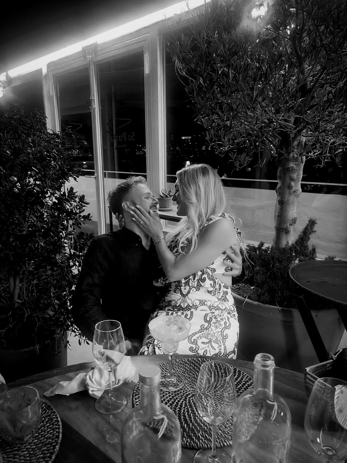 A couple holds hands, standing on a rocky beach under a large cave arch, gazing at the tranquil ocean during sunset. The sky is softly lit in pastel hues.