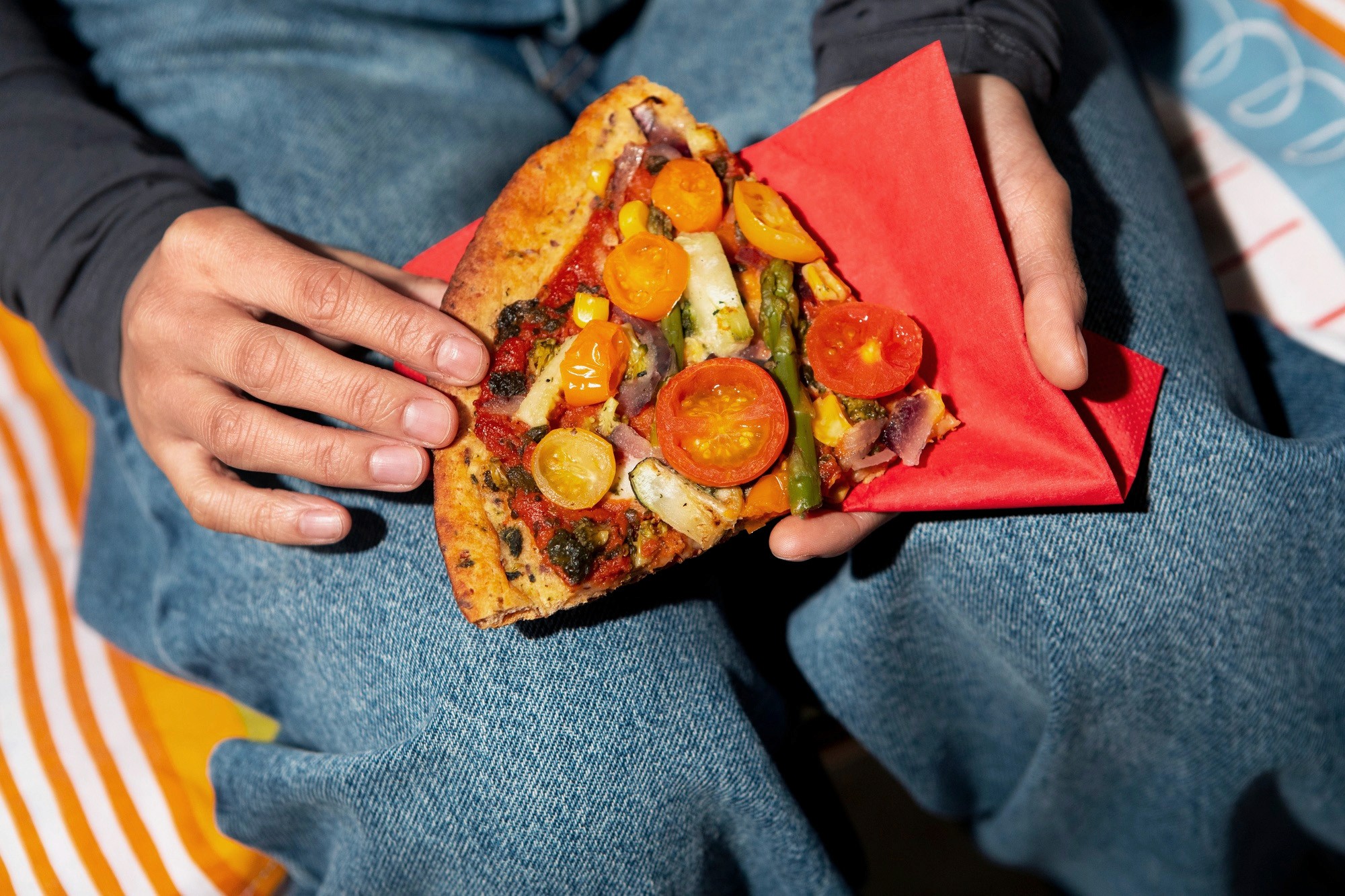 Close-up of a person's hands resting on their blue jeans, holding a slice of vegetable pizza on a red napkin.