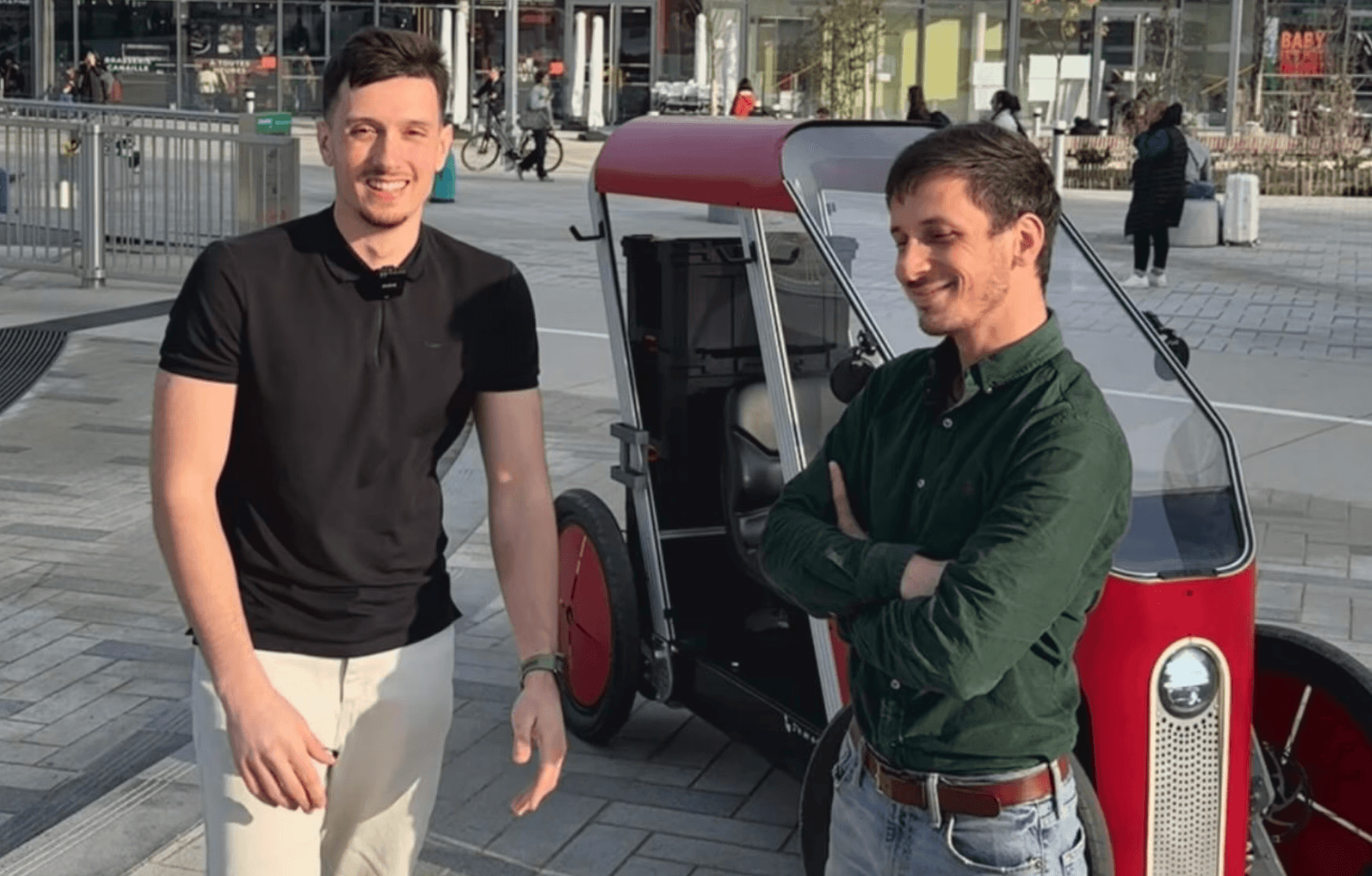 Two men standing next to a small red electric microcar, showing Charging Stack's hands-on mobility industry engagement.