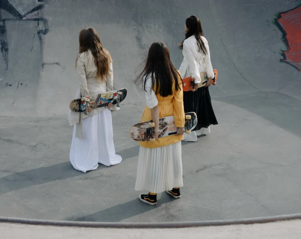 A group of young women wearing long skirts and flowing tops, each holding a skateboard, walk away from the camera in a concrete skatepark.