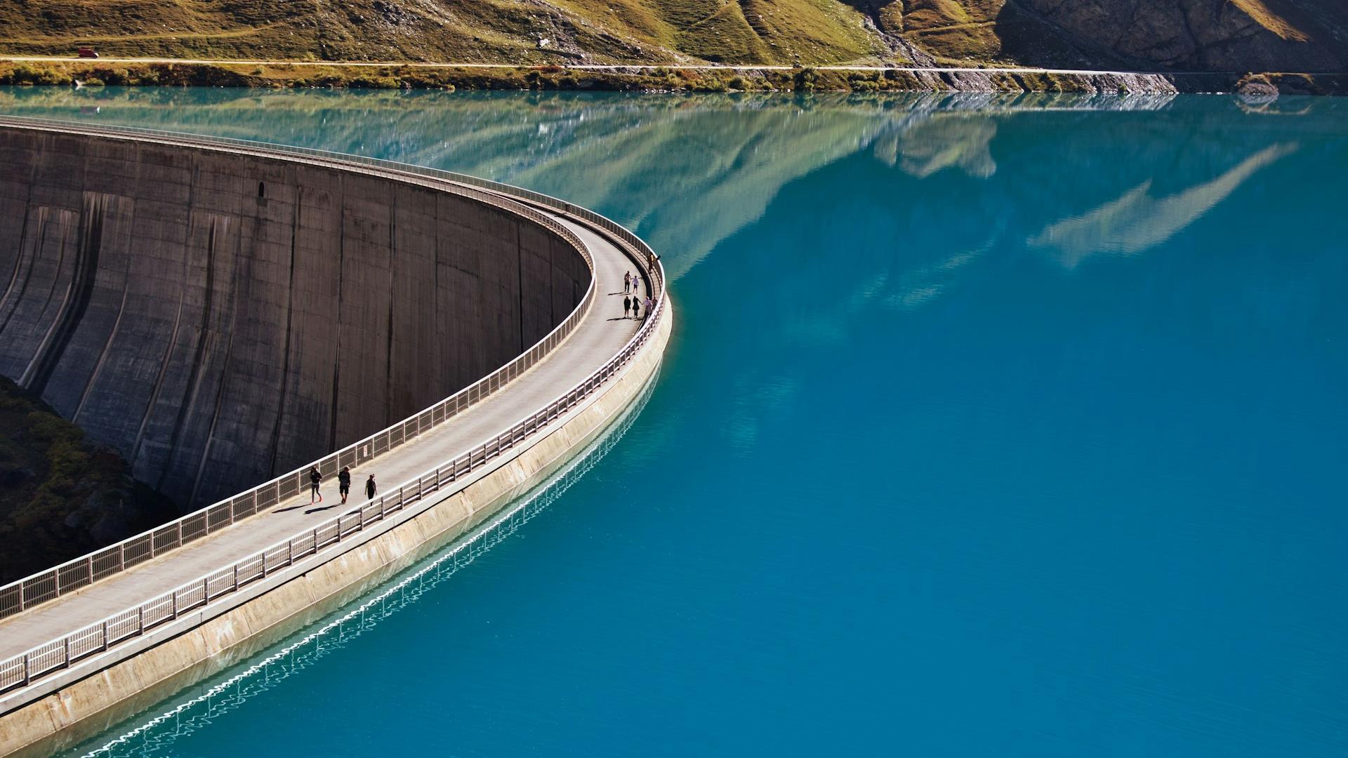 Curved concrete dam with people walking along it, set against a vibrant turquoise lake. Mountains are reflected in the calm water, creating a serene atmosphere.