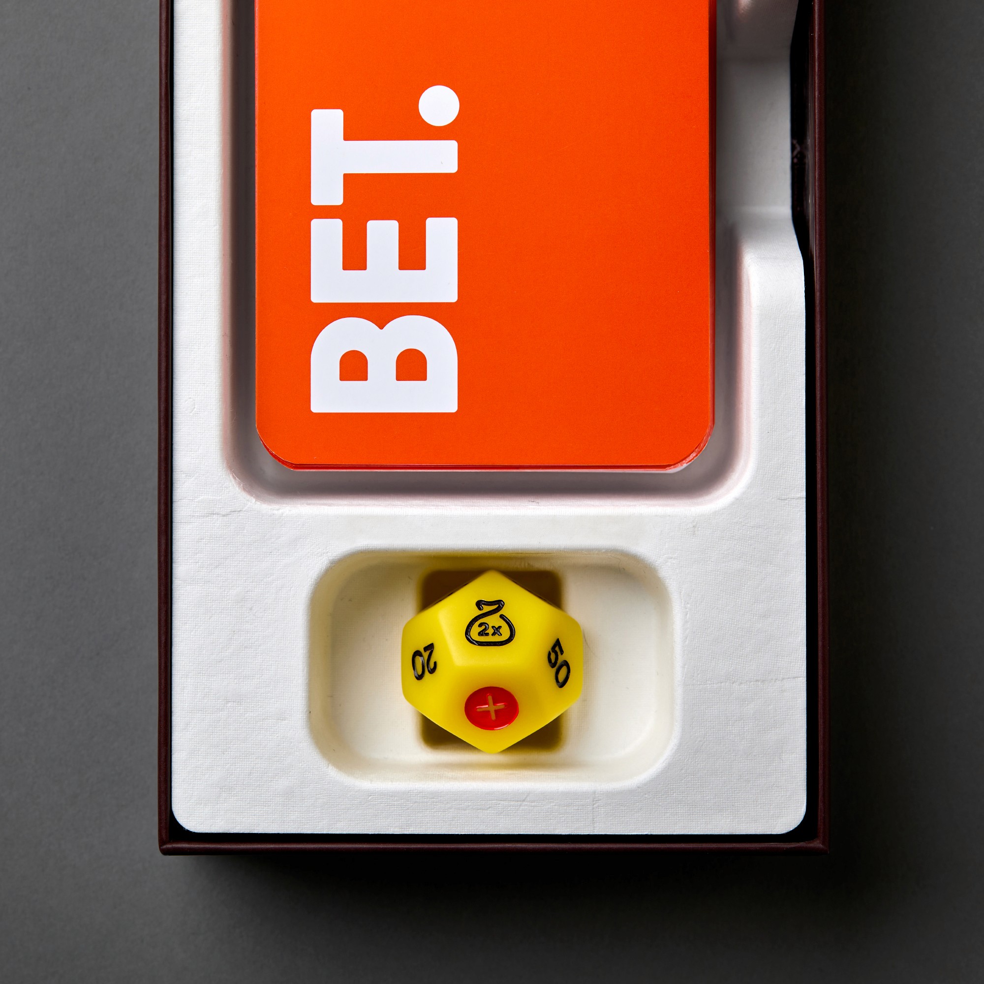 Overhead view of the open BET. box insert, showing the orange card deck and the yellow die nestled in a white molded pulp tray.