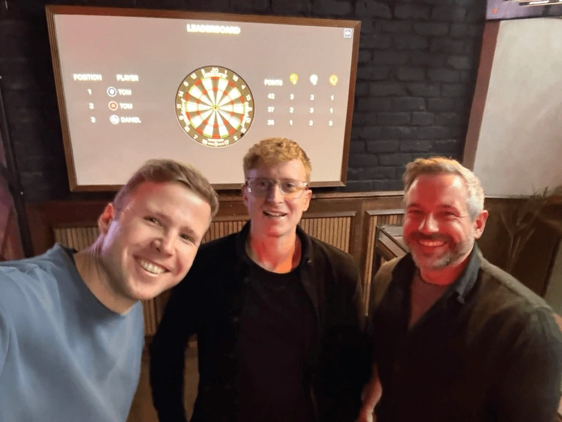 Team members posing in front of a darts leaderboard.