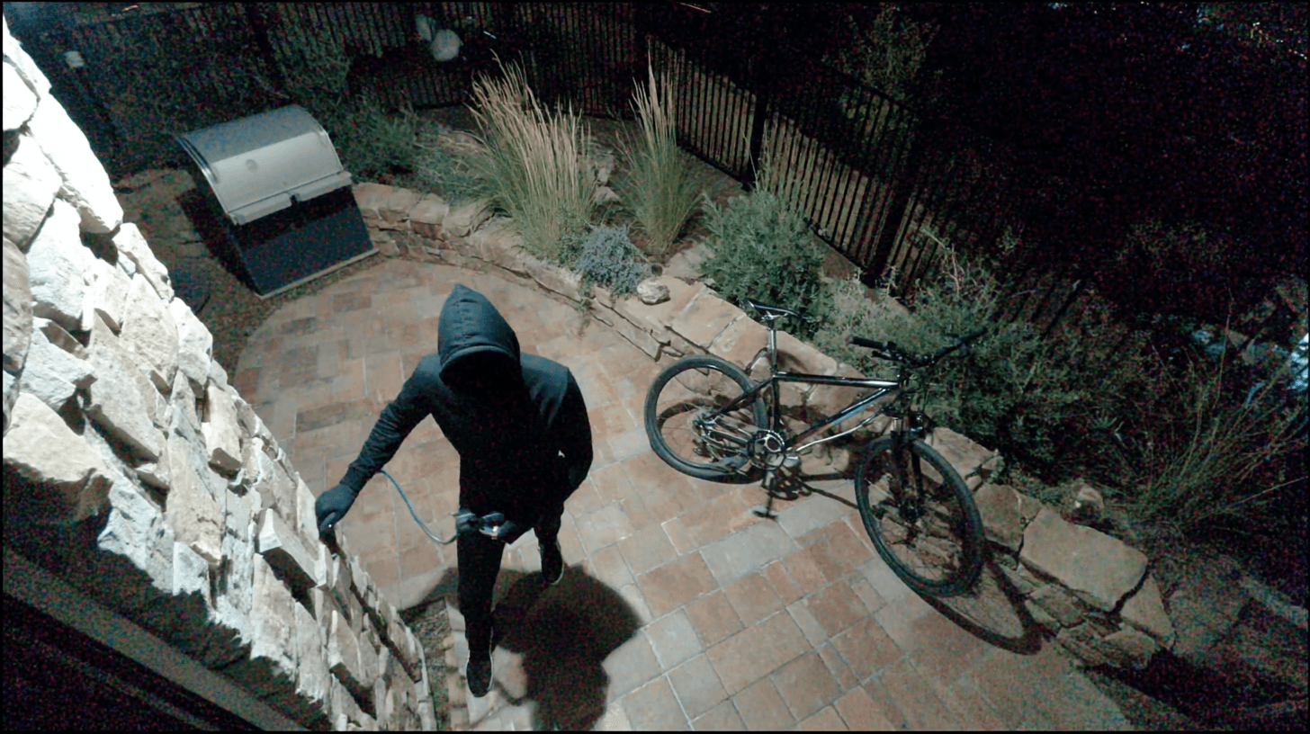 A person in a hooded jacket stands suspiciously near a brick patio at night, with a bicycle nearby, suggesting potential burglary activity.