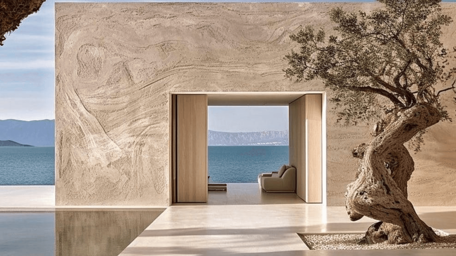 Minimalist stone residence entrance framing a sea view beside an old olive tree
