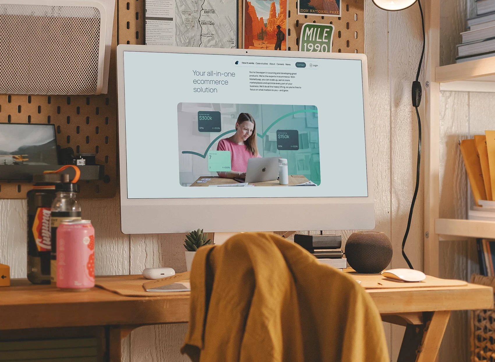 “Desktop monitor on a wooden desk displaying the MarketLeap website homepage. The website headline reads ‘Your all-in-one ecommerce solution’. A photo on the screen shows a woman using a laptop surrounded by data visualisations, including purchase values of ‘$300k’ and ‘$150k’. The workspace is homely and creative, featuring a pegboard wall, books, drinks, and a yellow jacket draped over the chair.”