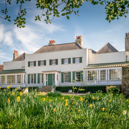 A white, two-story house with green shutters, surrounded by blooming flowers and greenery, viewed from the garden.