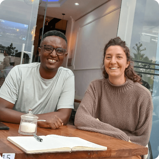 A young person with curly hair smiles at the camera while sitting in front of a laptop against a dark background.