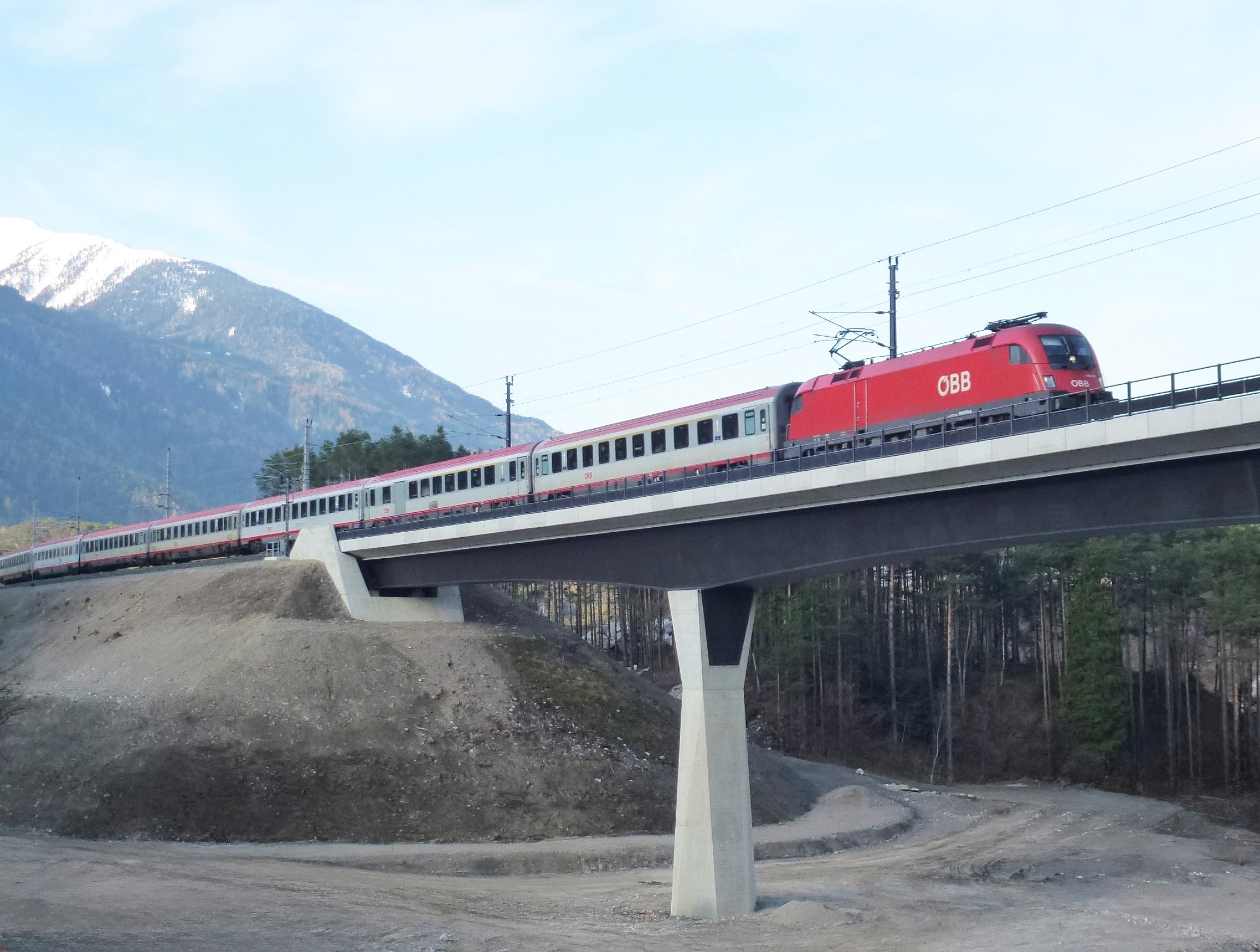 Roter Zug auf einer Betonbrücke mit Oberleitung, im Hintergrund bewaldete Hänge und schneebedeckter Berg.