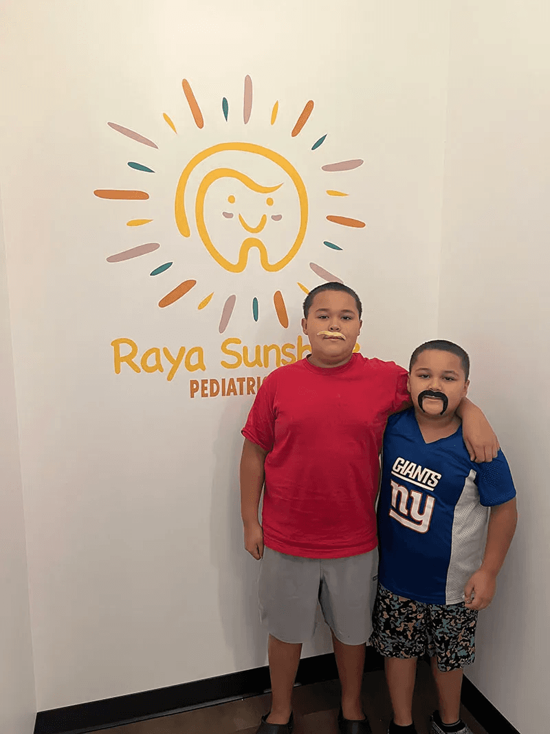 Two children standing side by side in front of the Raya Sunshine Pediatric Dentistry logo