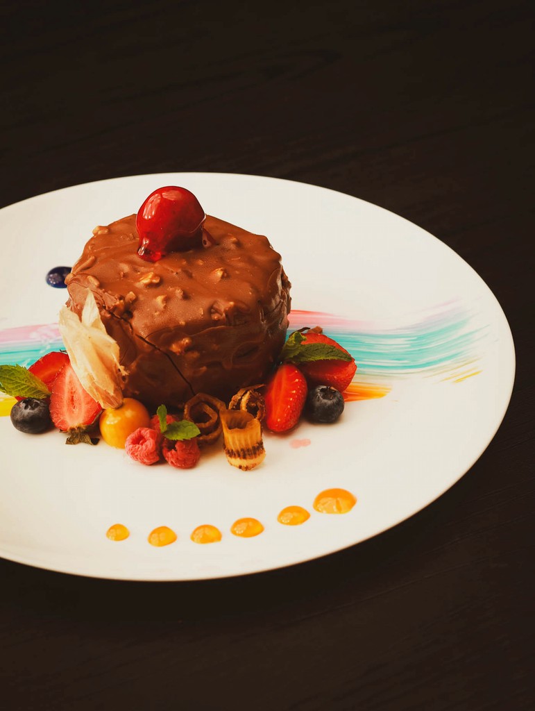 Gourmet chocolate dessert with nuts, fresh berries, and a cherry topping, elegantly plated on a white dish.