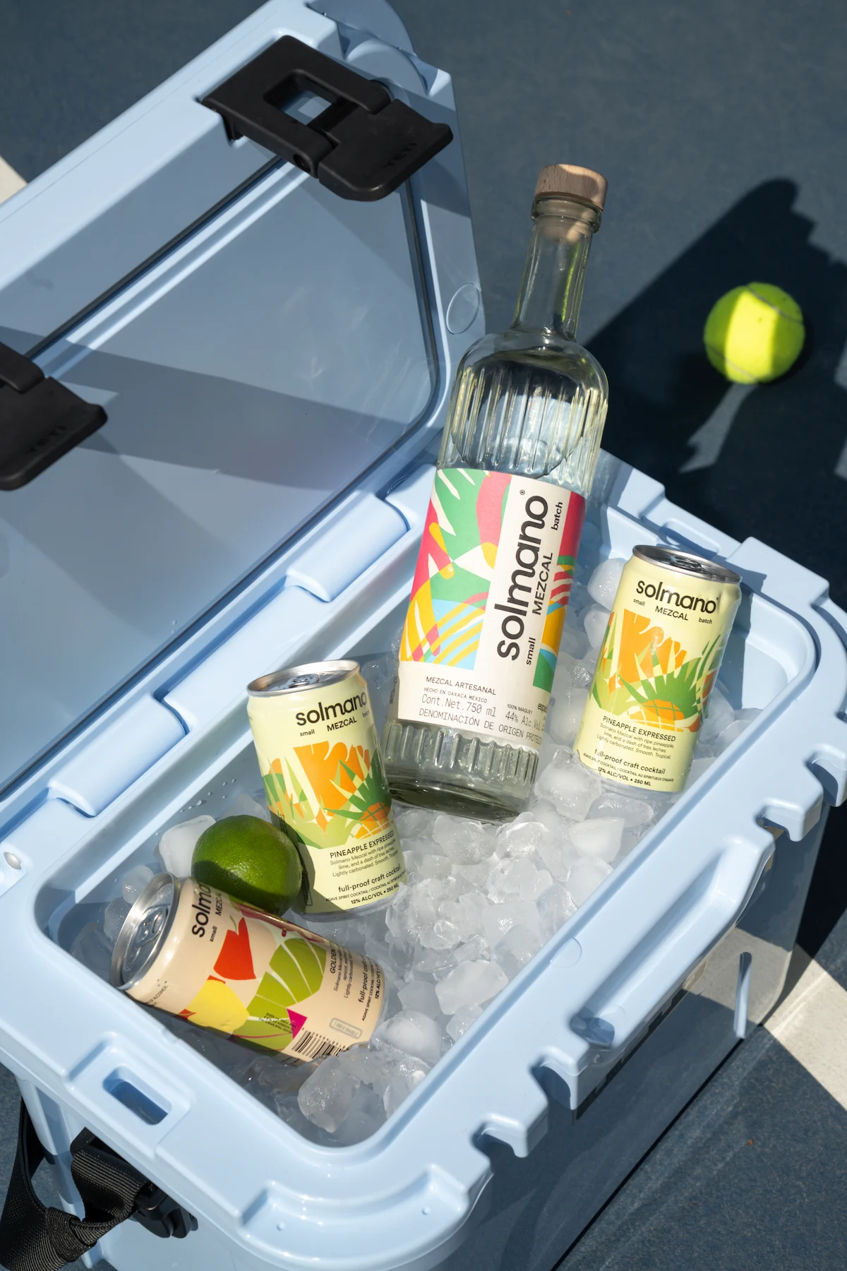 Solmano mezcal bottle and ready-to-serve cocktail cans displayed on marble beside fresh citrus and a pink cocktail against green tile