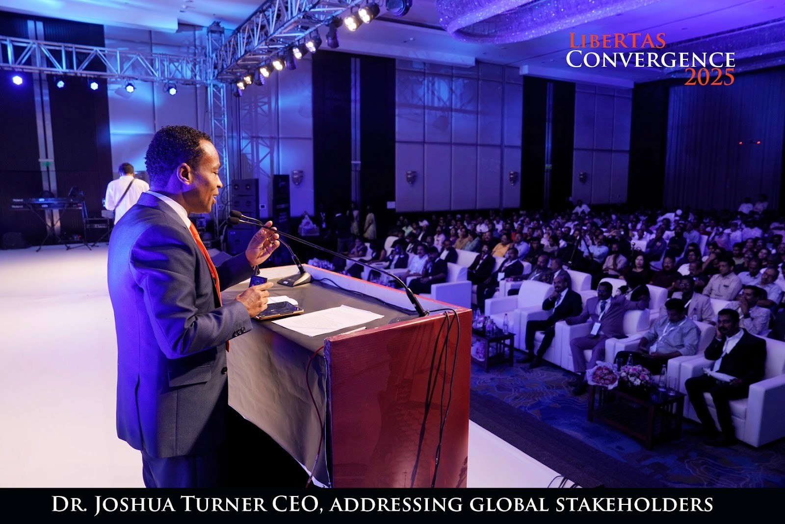 A speaker in a suit addresses a large audience at a conference. The setting is formal with blue lighting, emphasizing a professional atmosphere.