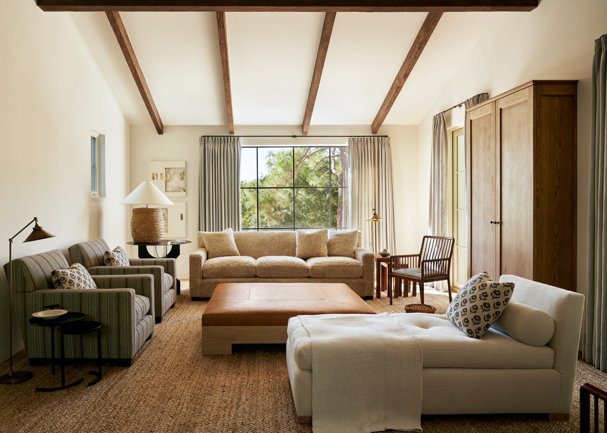 A living room with a bouclé sofa, green-striped armchairs, and a leather-topped coffee table on a jute rug. Exposed wood beams cross the ceiling above, and natural light filters through a steel-frame window framed by linen curtains.