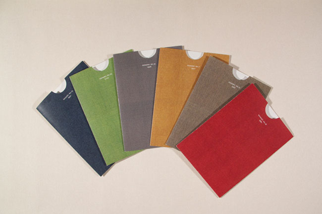The image shows a fan of six rectangular cards arranged on a white background. The cards are slightly curved and overlap each other, creating a sense of depth. Each card has a white circular cutout at the top center and a small white label with text printed in a light color, likely indicating the brand or product name. The cards are colored in muted earthy tones: dark blue, green, gray, orange-brown, and red.