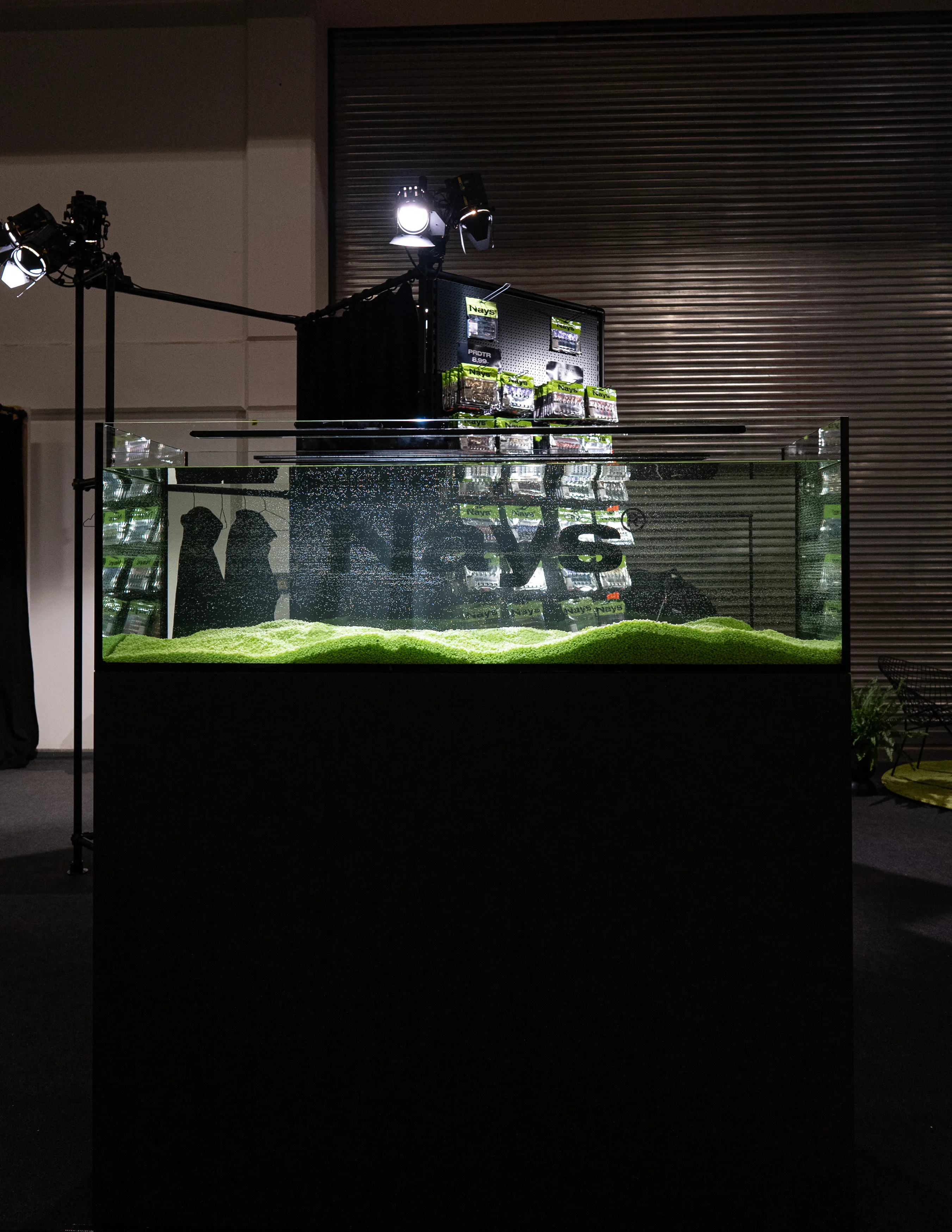 Spotlit aquarium with green gravel and Nays lettering on the glass at the dark Nays trade fair booth.