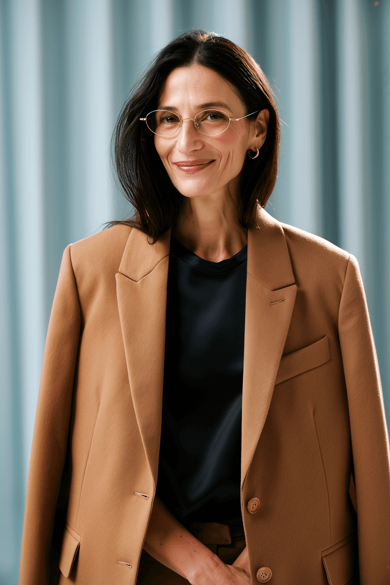 Smiling woman with long dark hair and glasses, wearing a tan blazer over a black shirt. Stands against a soft blue background, conveying confidence.