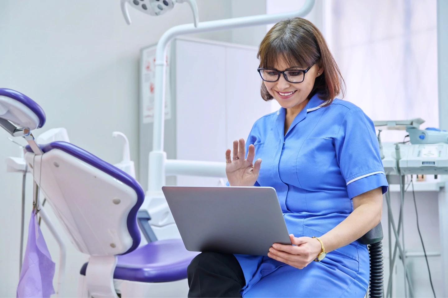 A dental professional in blue scrubs sits in a modern dental office, engaging in a video call on a laptop while a dental chair and equipment are visible in the background.
