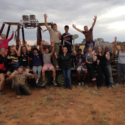 A group of people posing happily with raised hands in front of a hot air balloon basket in a dry, grassy field.