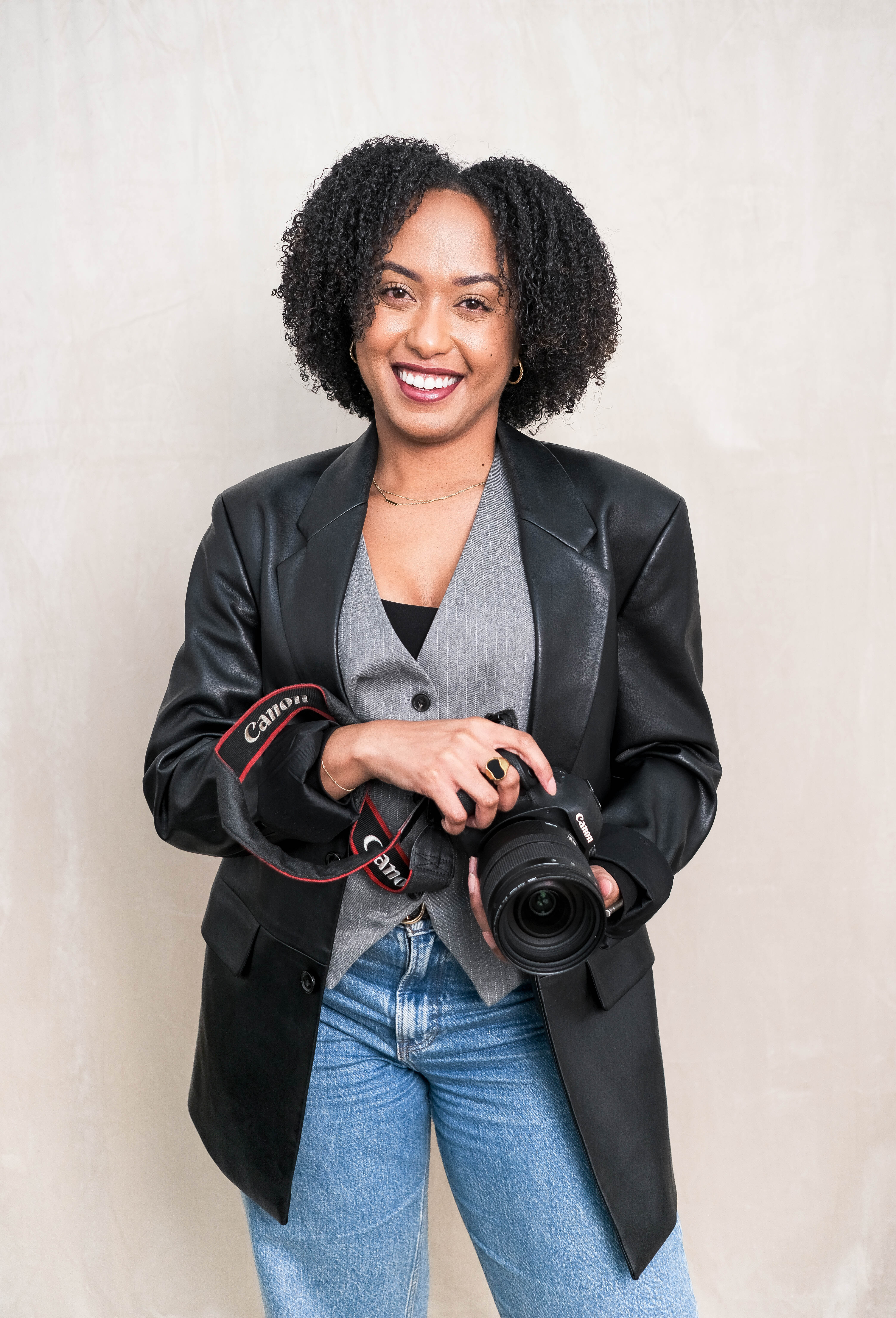 A woman with porcelain skin, striking freckles, and red lips poses against a dark background. She wears a brown textured coat and looks confidently at the camera, with vibrant orange flowers adding warmth to the composition.