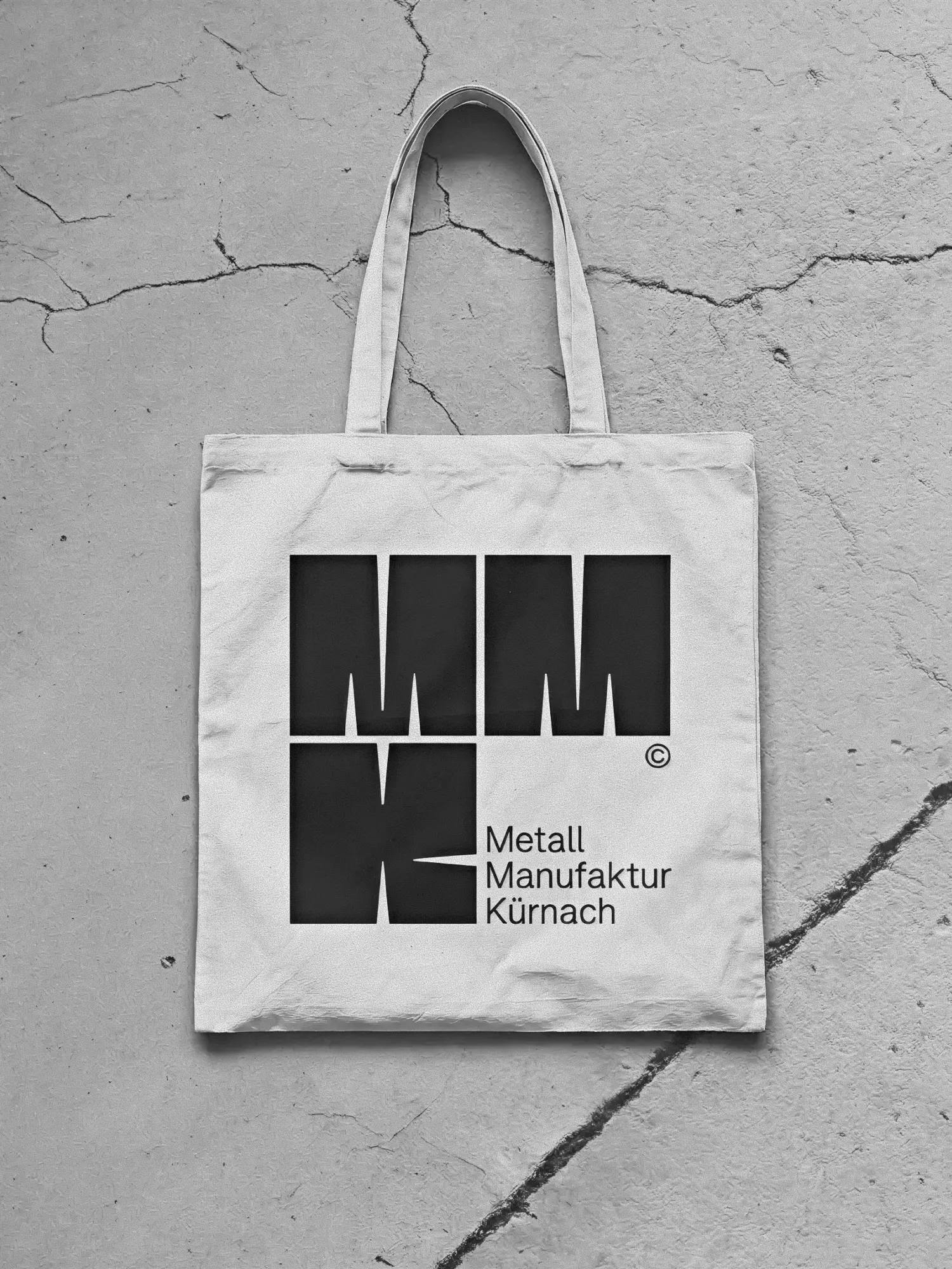 Weiße Stofftasche mit schwarzem MMK-Logo auf rissigem Betonboden, Schwarzweiß-Aufnahme