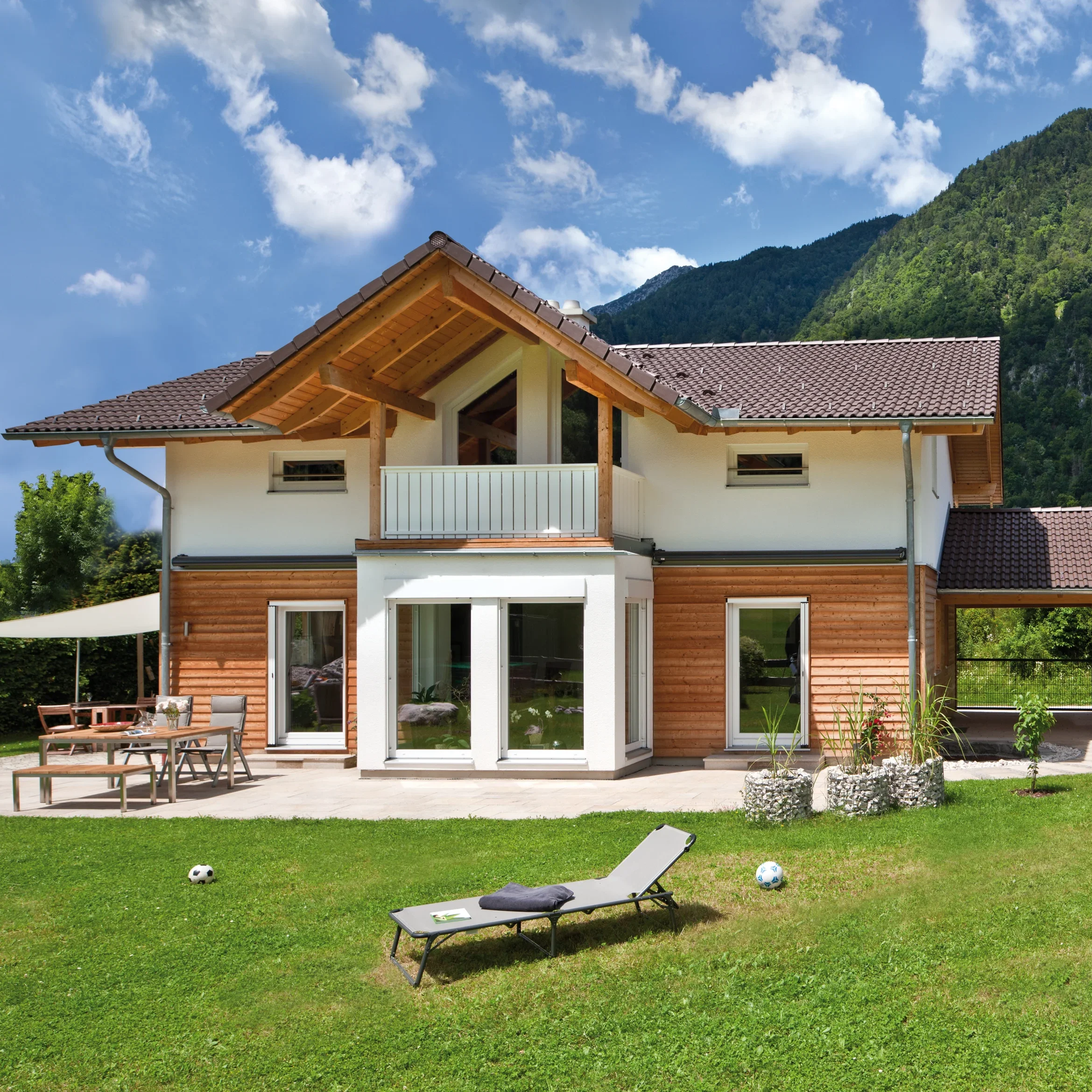 Modernes Holzhaus mit großer Terrasse und Garten, eingebettet in eine Berglandschaft unter blauem Himmel.