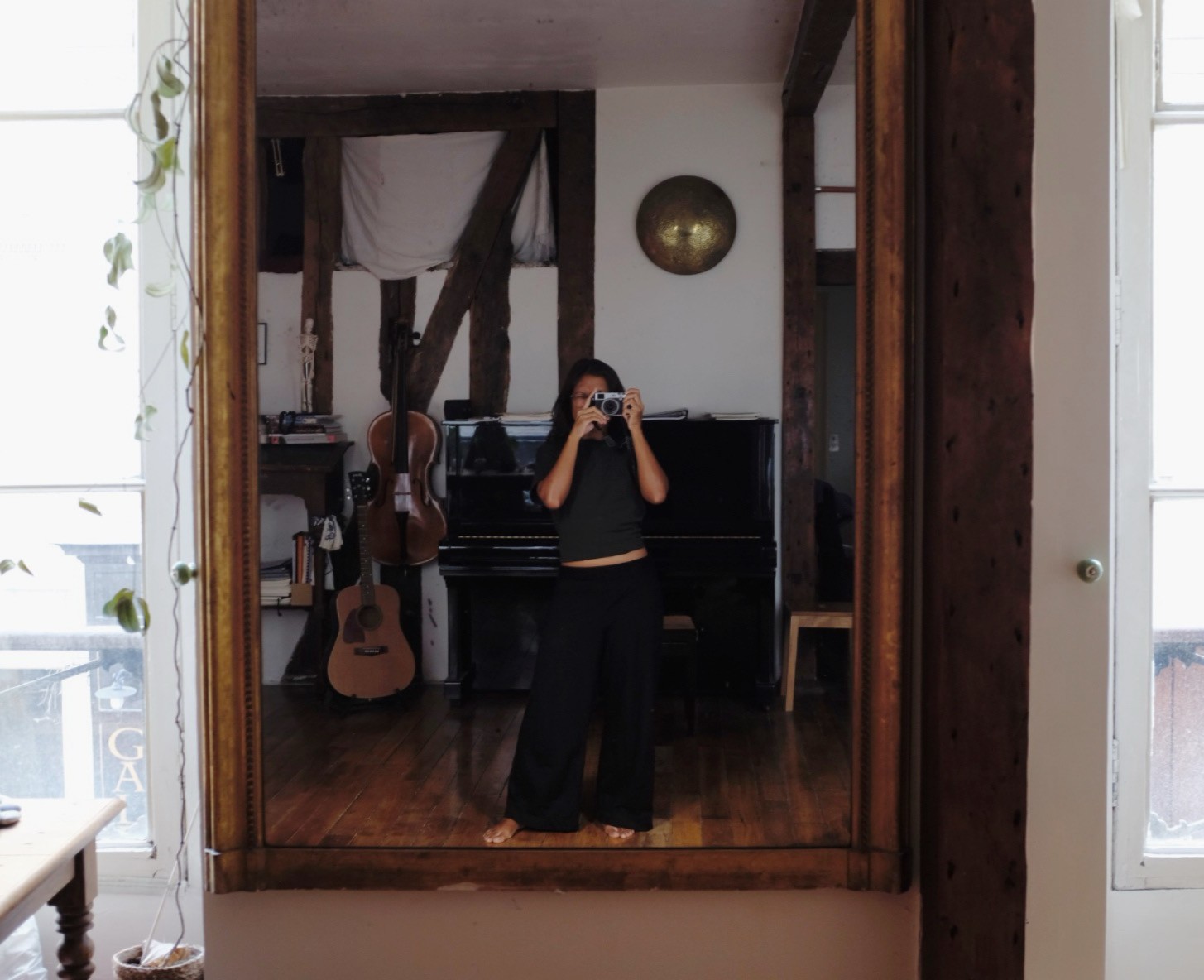 Una persona con una camiseta negra y pantalones de color claro está de pie en la cima rocosa de una montaña al atardecer, sosteniendo una cámara frente a su cara para tomar una foto. Un mar de nubes se extiende detrás de ellos, iluminado por la luz dorada del sol poniente.