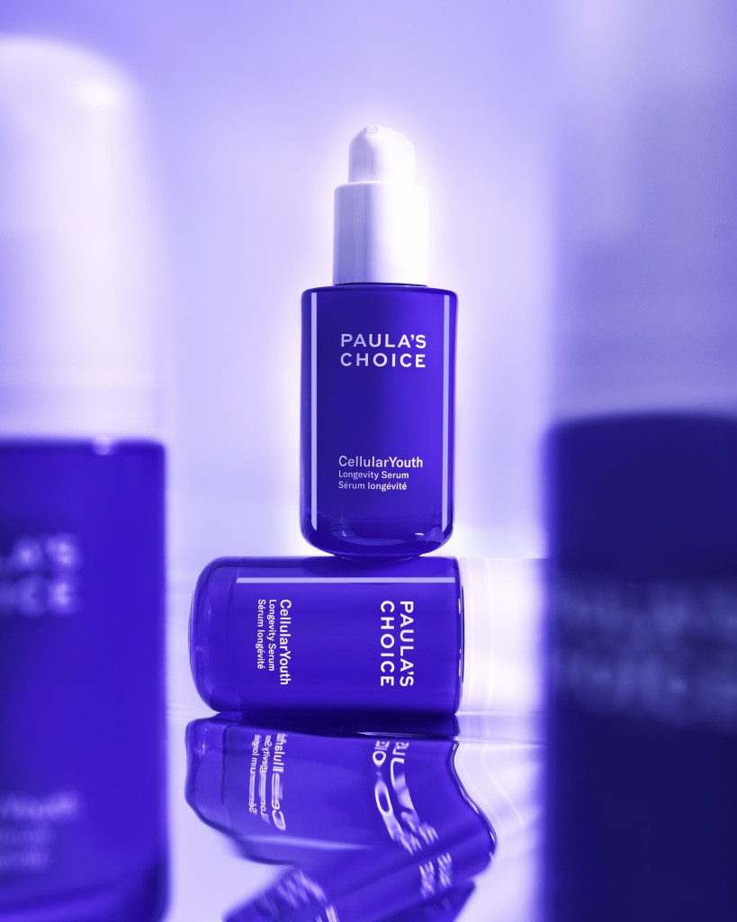 Two bottles of Paula's Choice Cellular Youth Longevity Serum in a purple hue, with one bottle upright and one lying down, reflecting on a smooth surface.