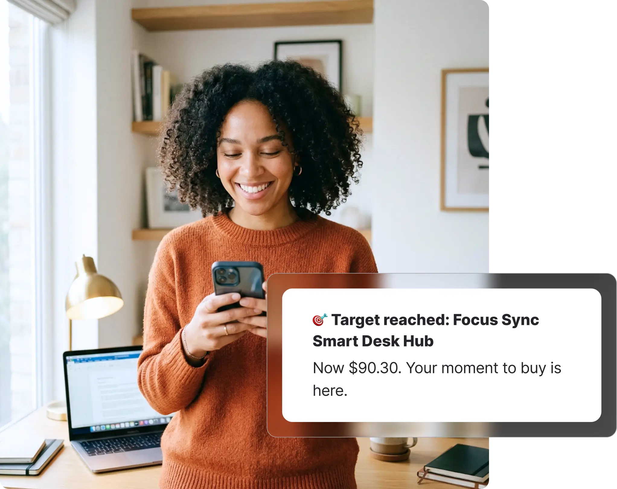 Woman with curly hair smiling at her phone in a bright home office, receiving a JB Hi-Fi target price notification on Whisprice, with a laptop and books on a desk beside her.