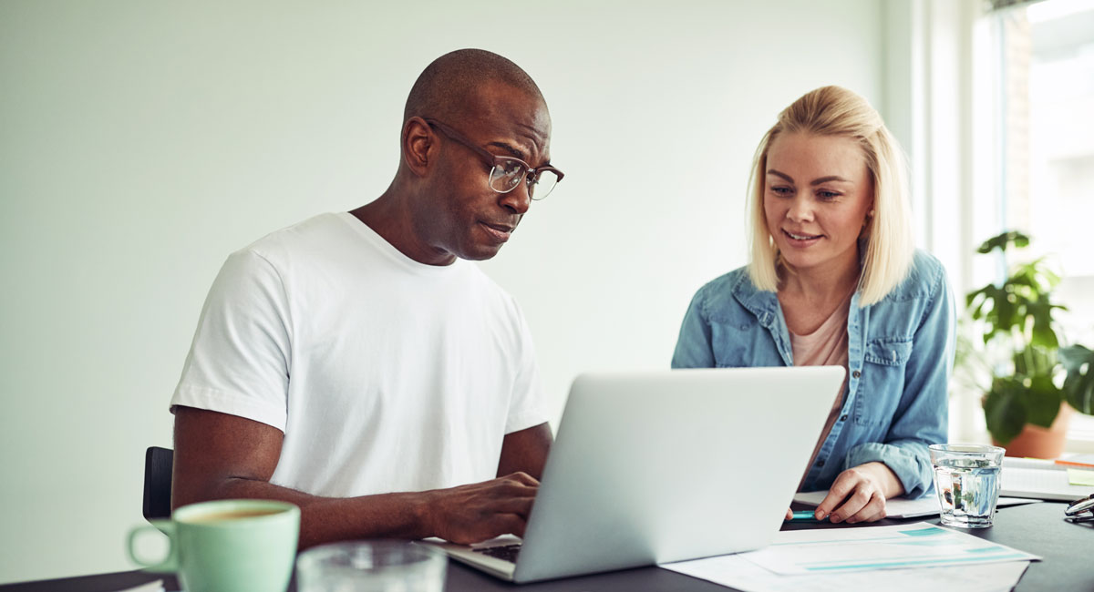 Two colleagues reviewing work together on a laptop at a desk with documents and a water glass