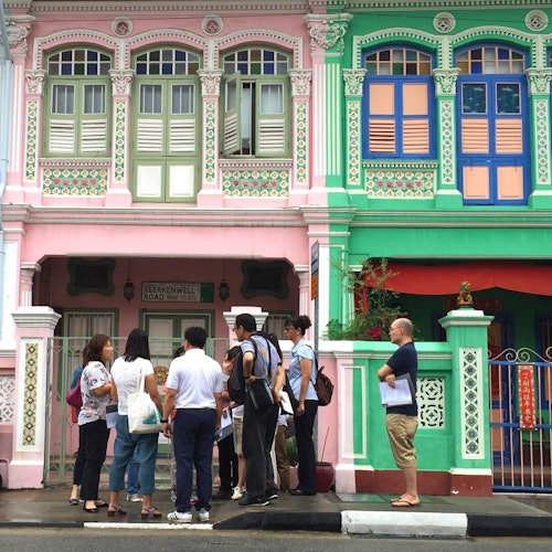 A group of people stands in front of colorful, ornately decorated buildings, with pink and green facades, and decorative windows.