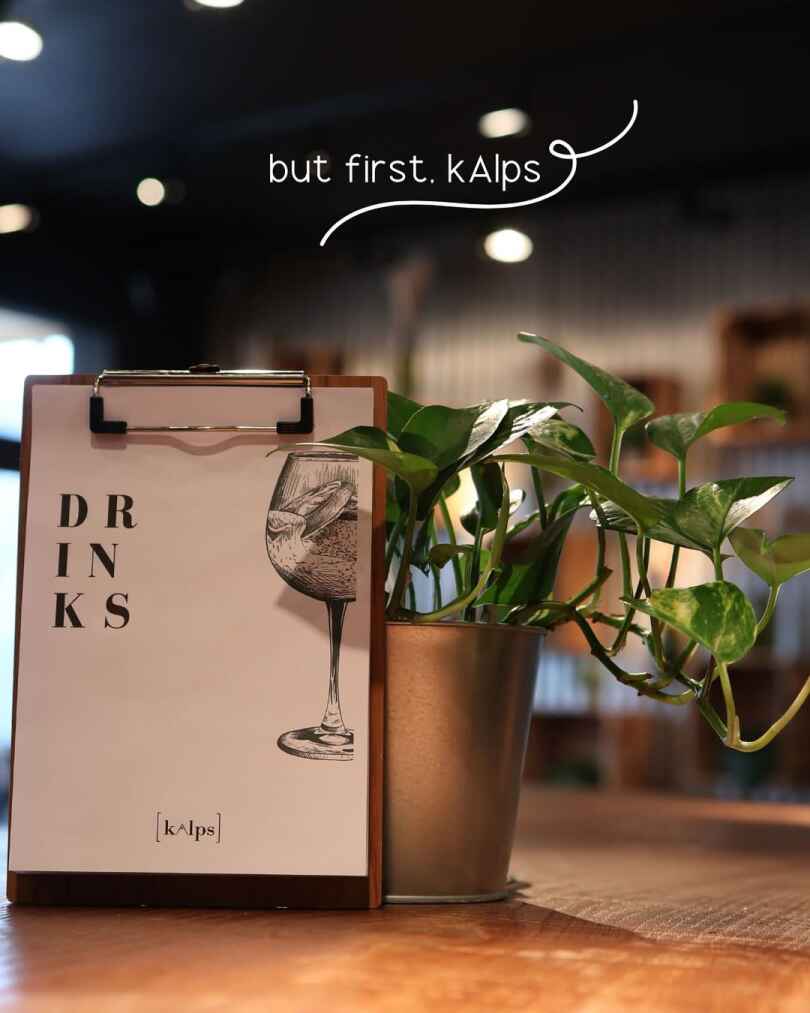 A drinks menu on a clipboard beside a potted plant on a wooden table inside a restaurant.