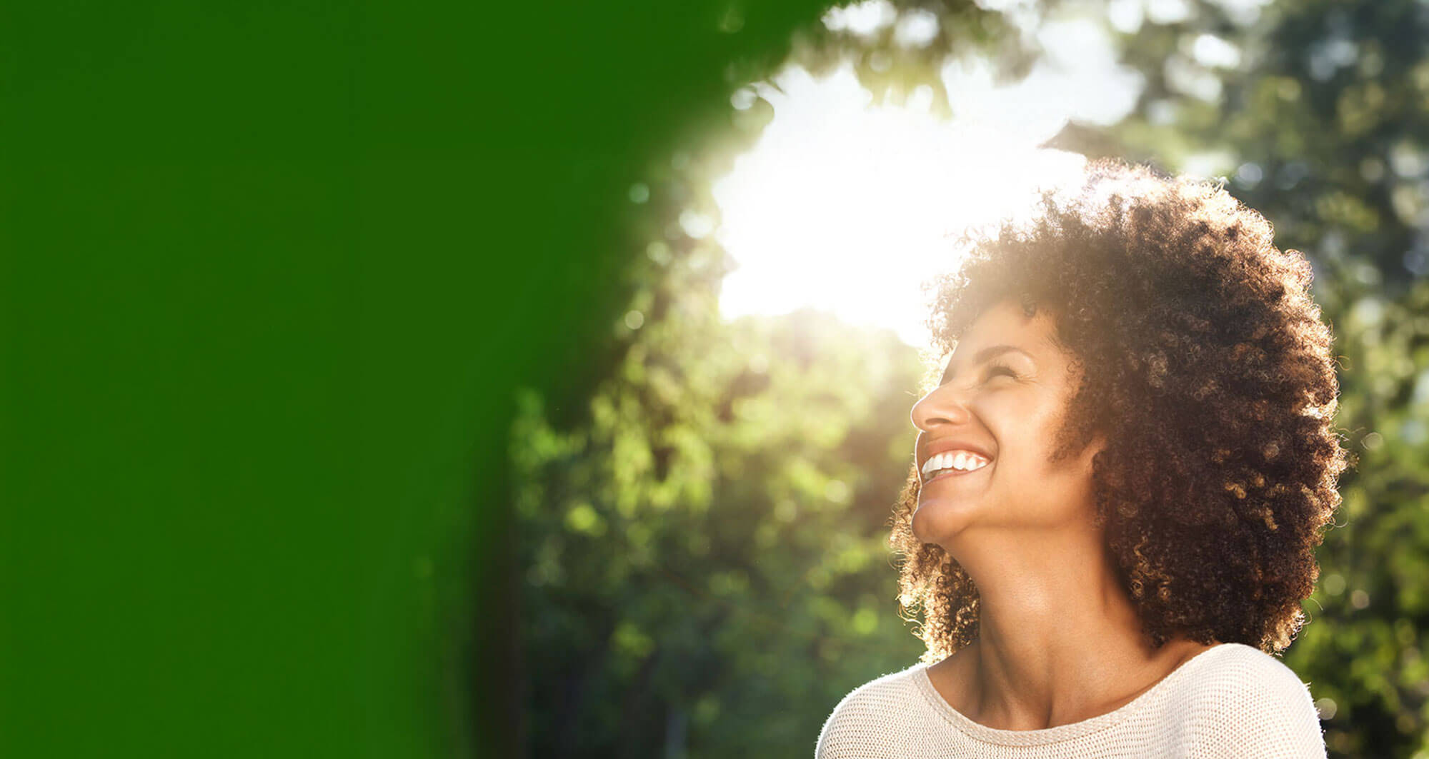 Smiling woman with curly hair enjoying sunshine in a lush green park.