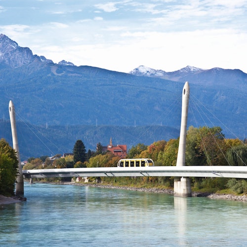 Uma moderna ponte estaiada com um bonde cruzando sobre um rio, tendo ao fundo árvores verdes, uma igreja e montanhas.