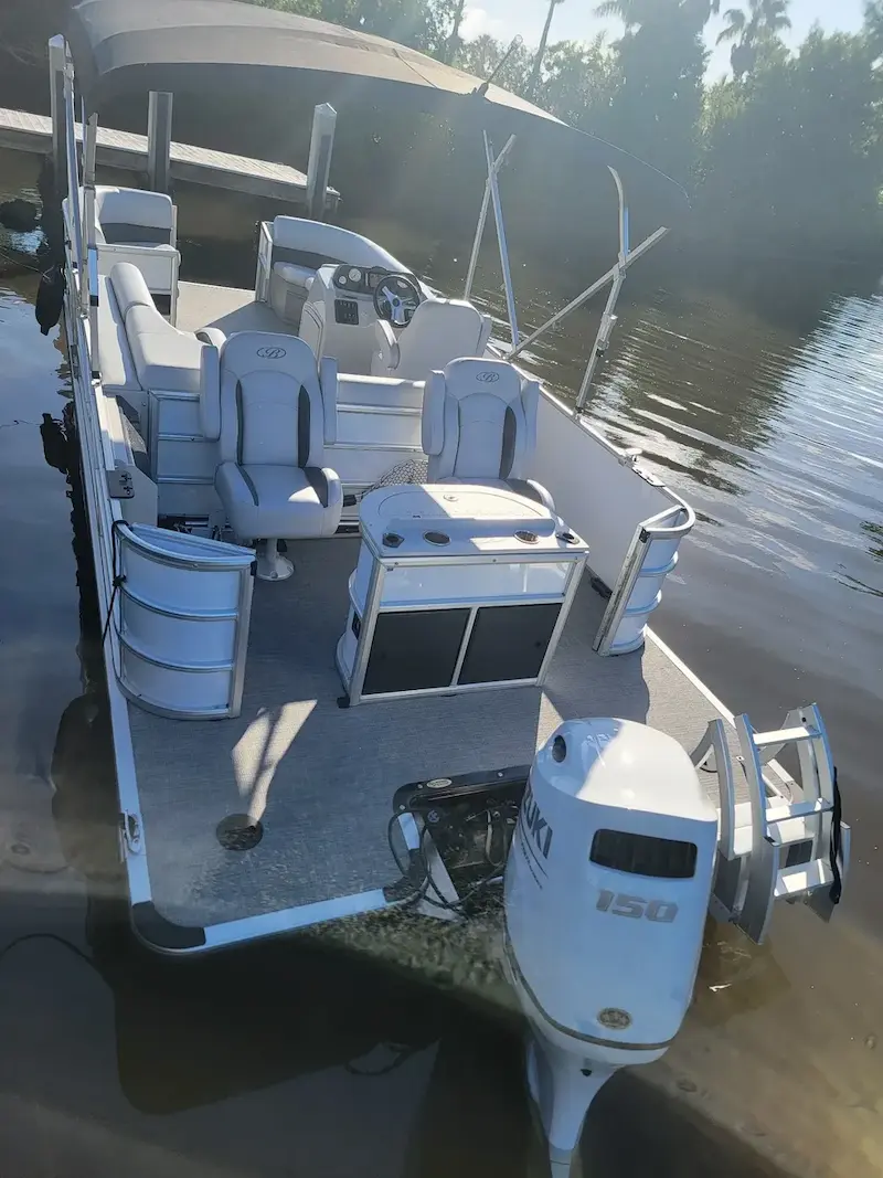 A white pontoon boat with comfortable seating and a powerful outboard motor is moored on a calm, reflective body of water, surrounded by lush greenery, capturing a serene boating scene in Bradenton, FL.