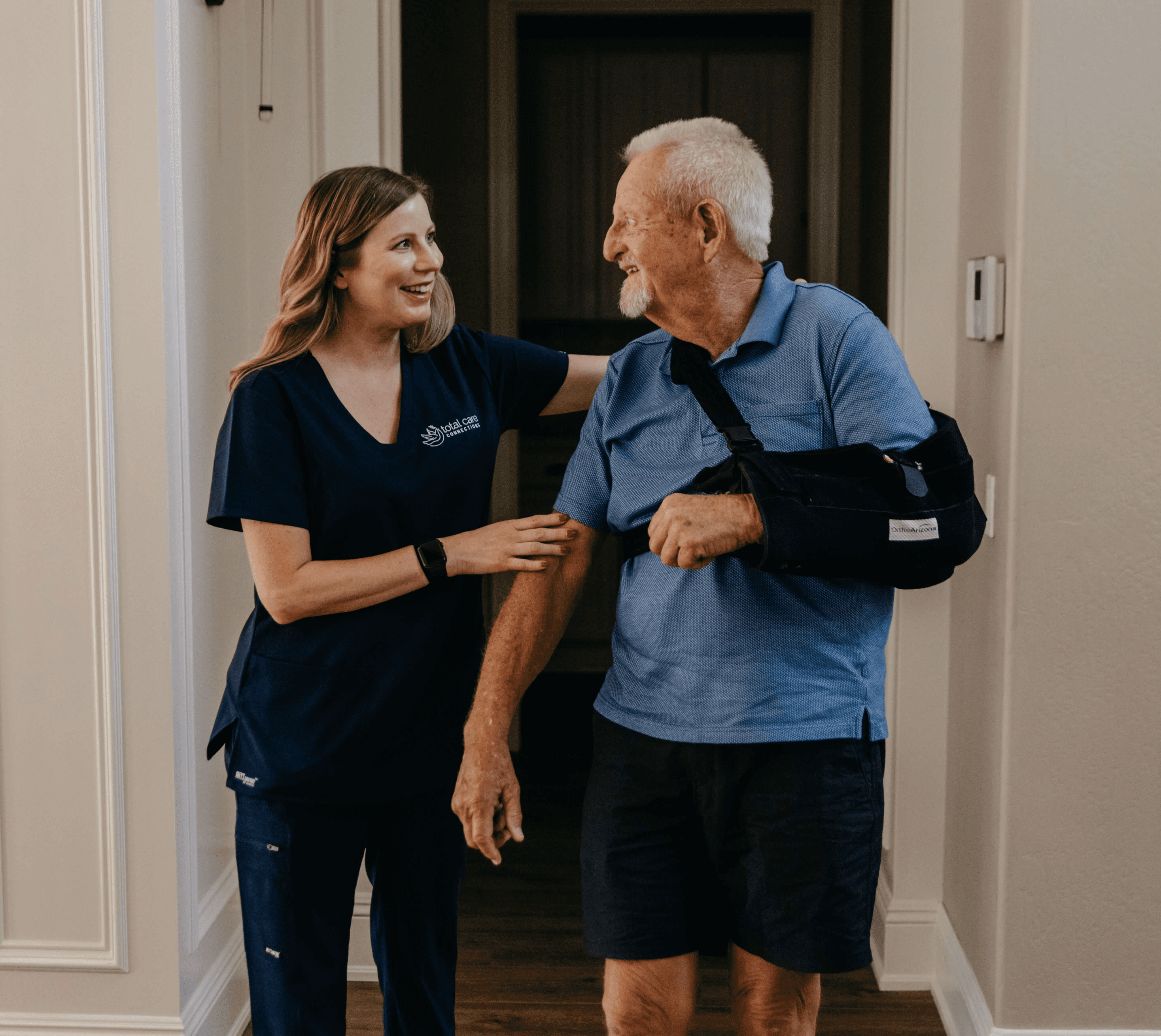 A woman in scrubs helps a smiling man with a sling on his arm walk through a doorway.