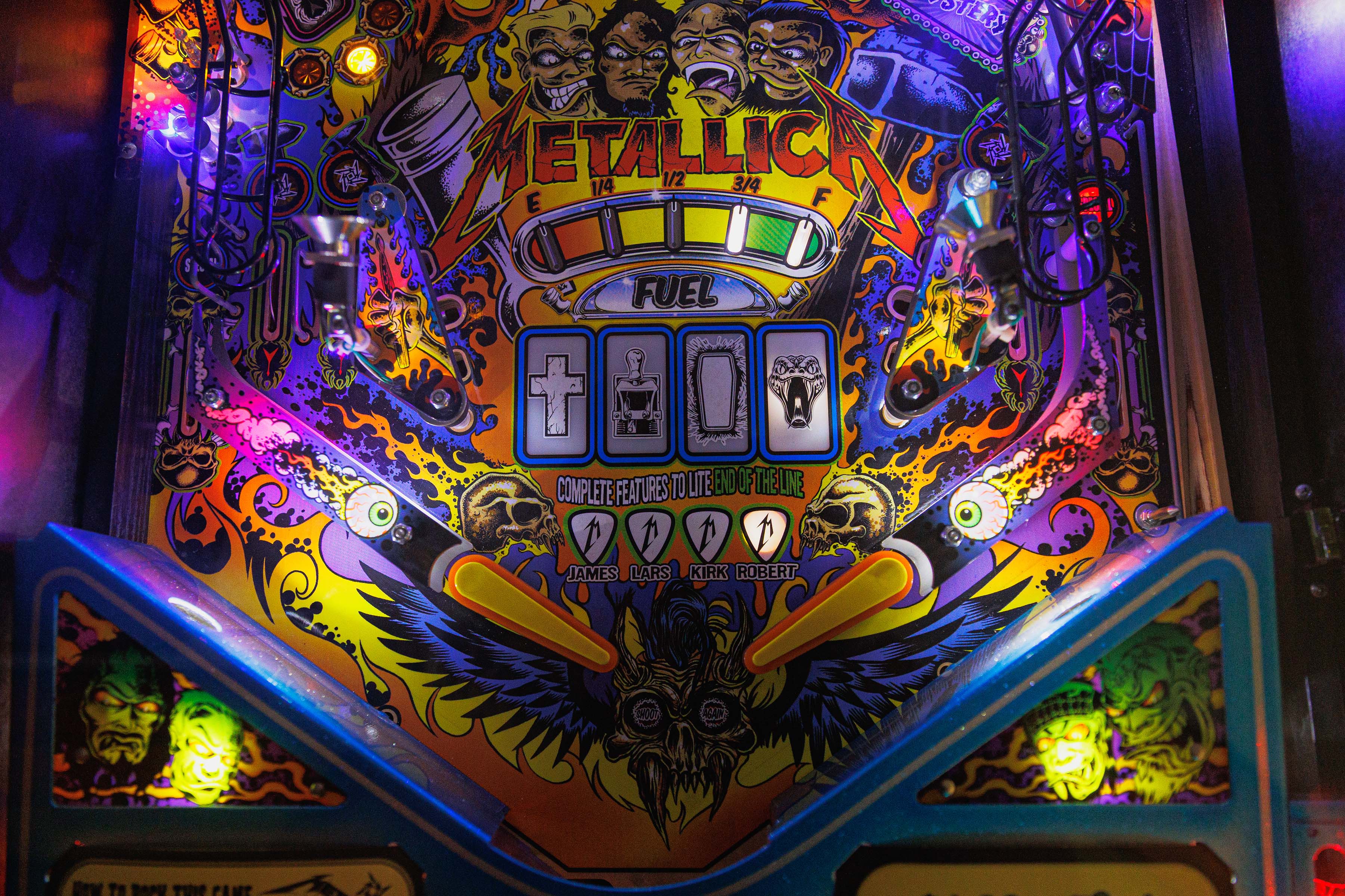 Close-up of a brightly lit Metallica pinball machine featuring vivid monster artwork, skulls, flames, and the band’s logo above targets labeled James, Lars, Kirk, and Robert.