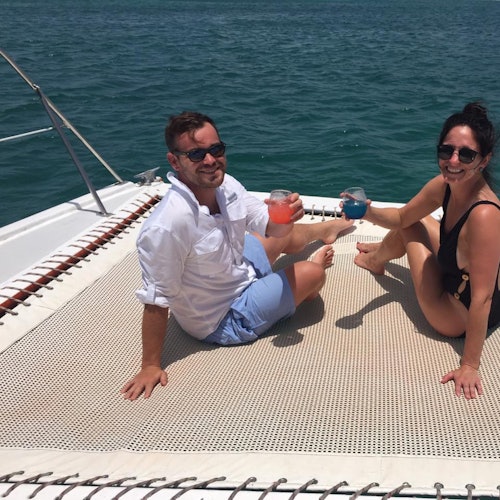 Two people sitting on a boat's netting, toasting drinks, smiling, and enjoying sunny weather with the ocean in the background.