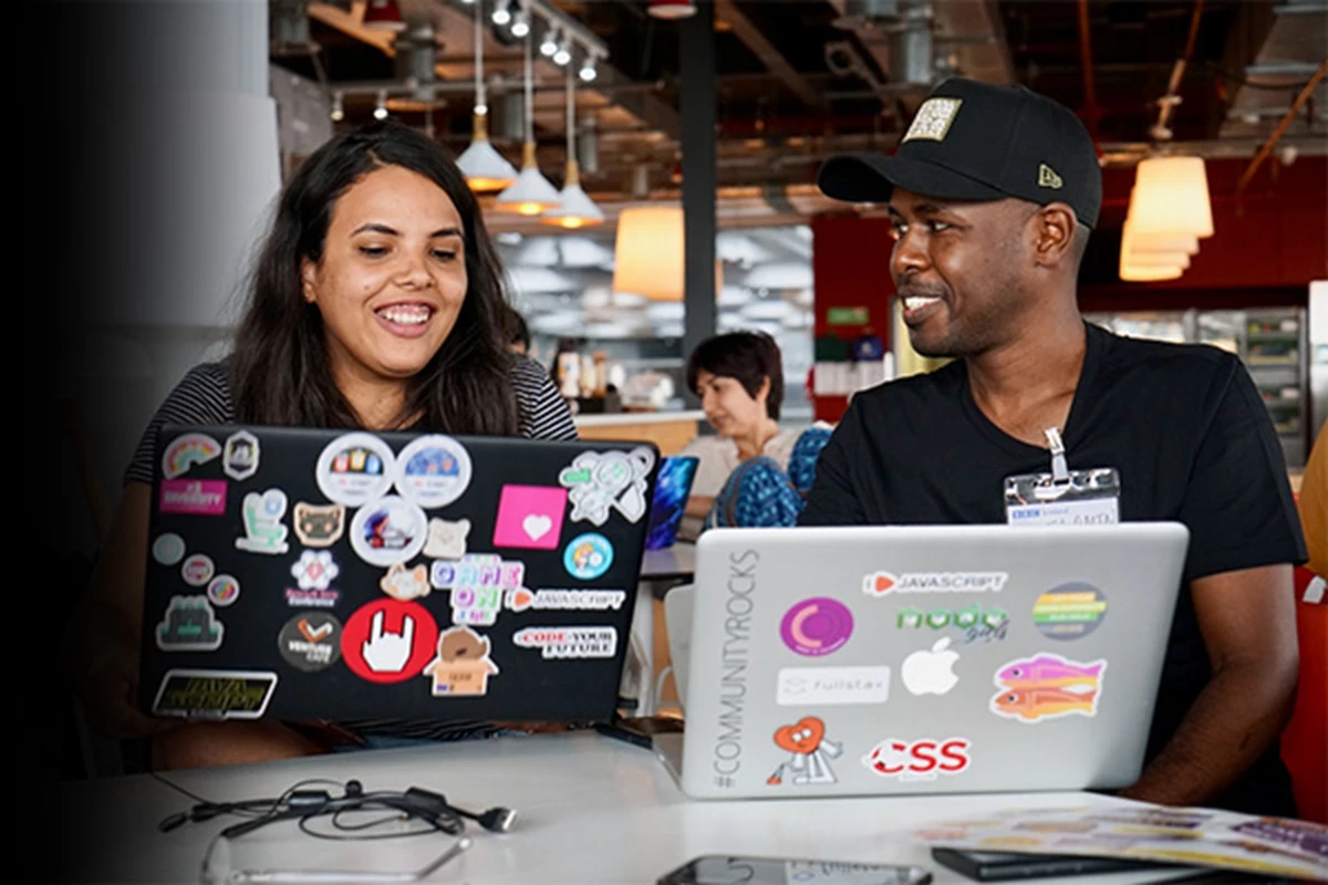 A woman and a man sat in front of laptops covered in stickers, they are smiling and laughing, the man is looking at the woman. They are in an open plan office with other people working on laptops in the background