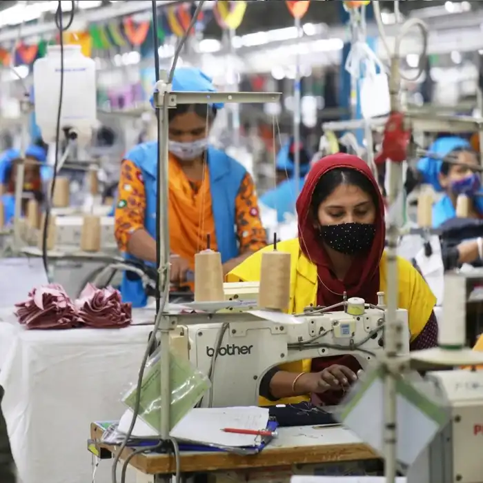 Female workers in a busy textile manufacturing unit using industrial sewing machines to produce clothing. Workers are wearing safety masks and uniforms, highlighting workplace safety and productivity in the garment industry.