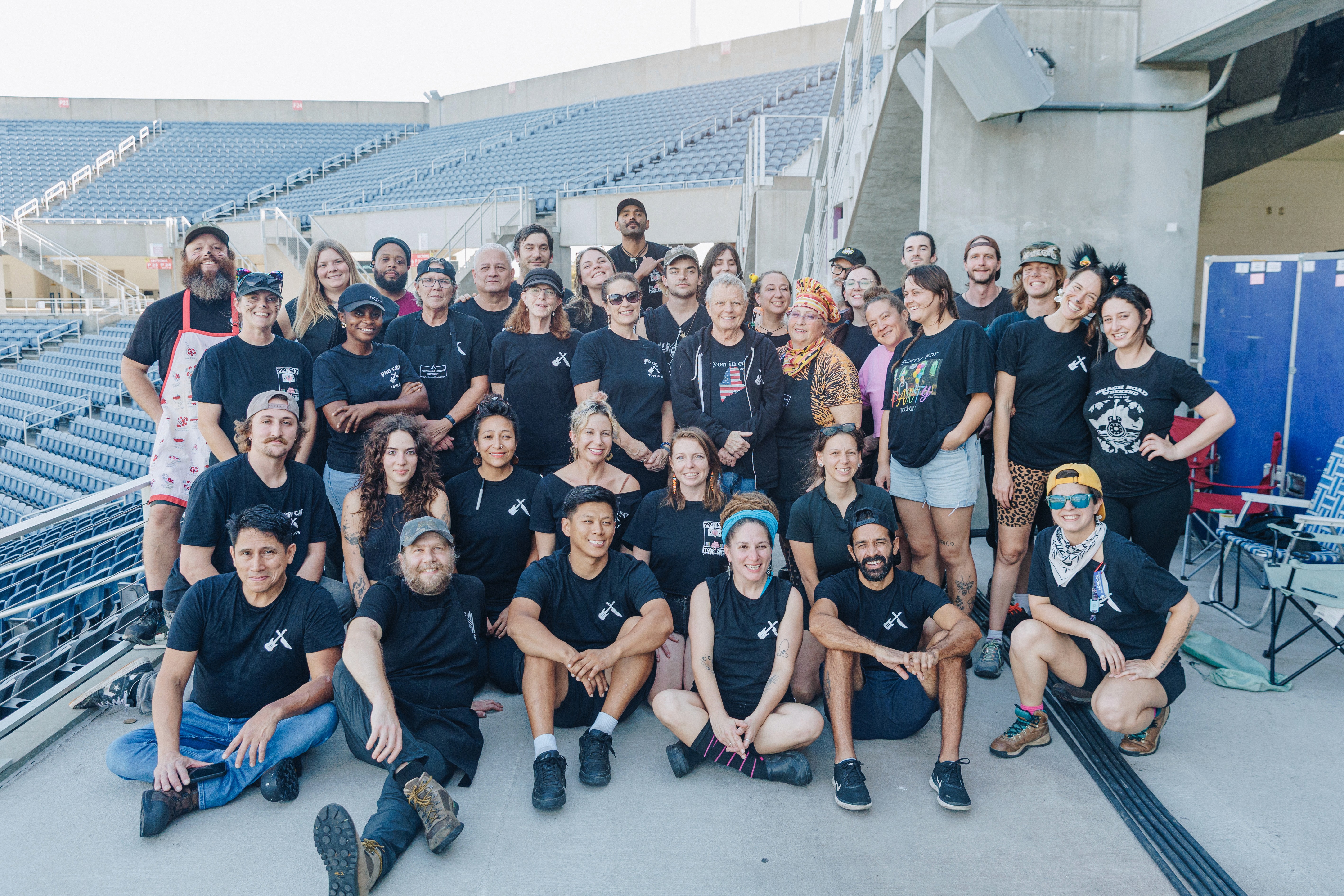 A large group of people in matching black shirts poses together outdoors, smiling and enjoying their time.