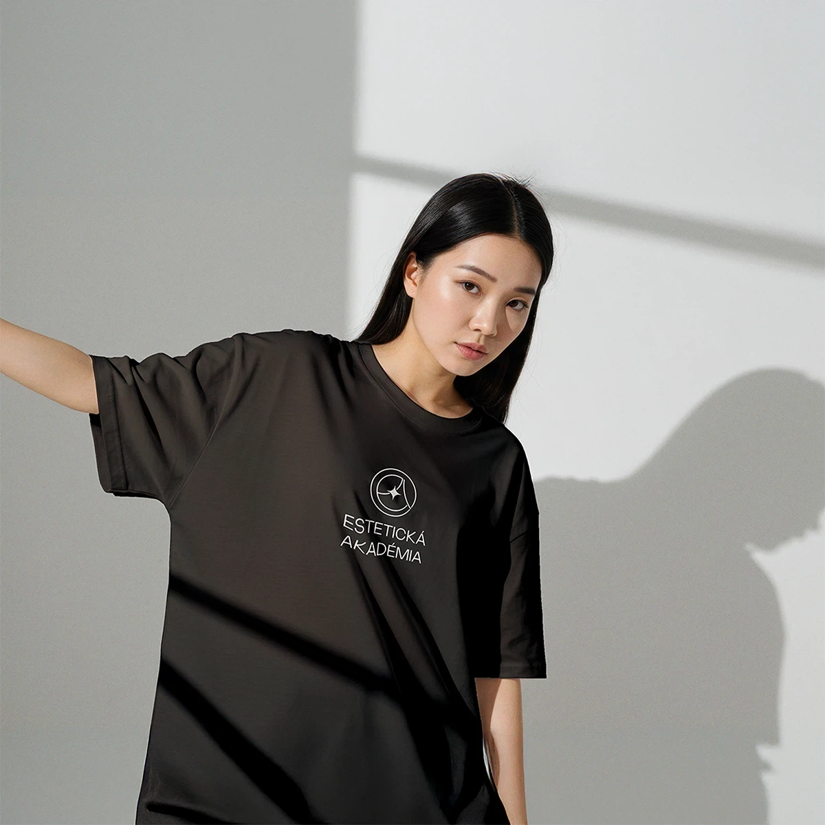 A person wearing a dark oversized shirt poses against a light wall, casting a shadow in soft natural light.