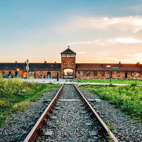 Railway tracks leading to a brick building with a central watchtower, surrounded by grassy fields under a partially cloudy sky.