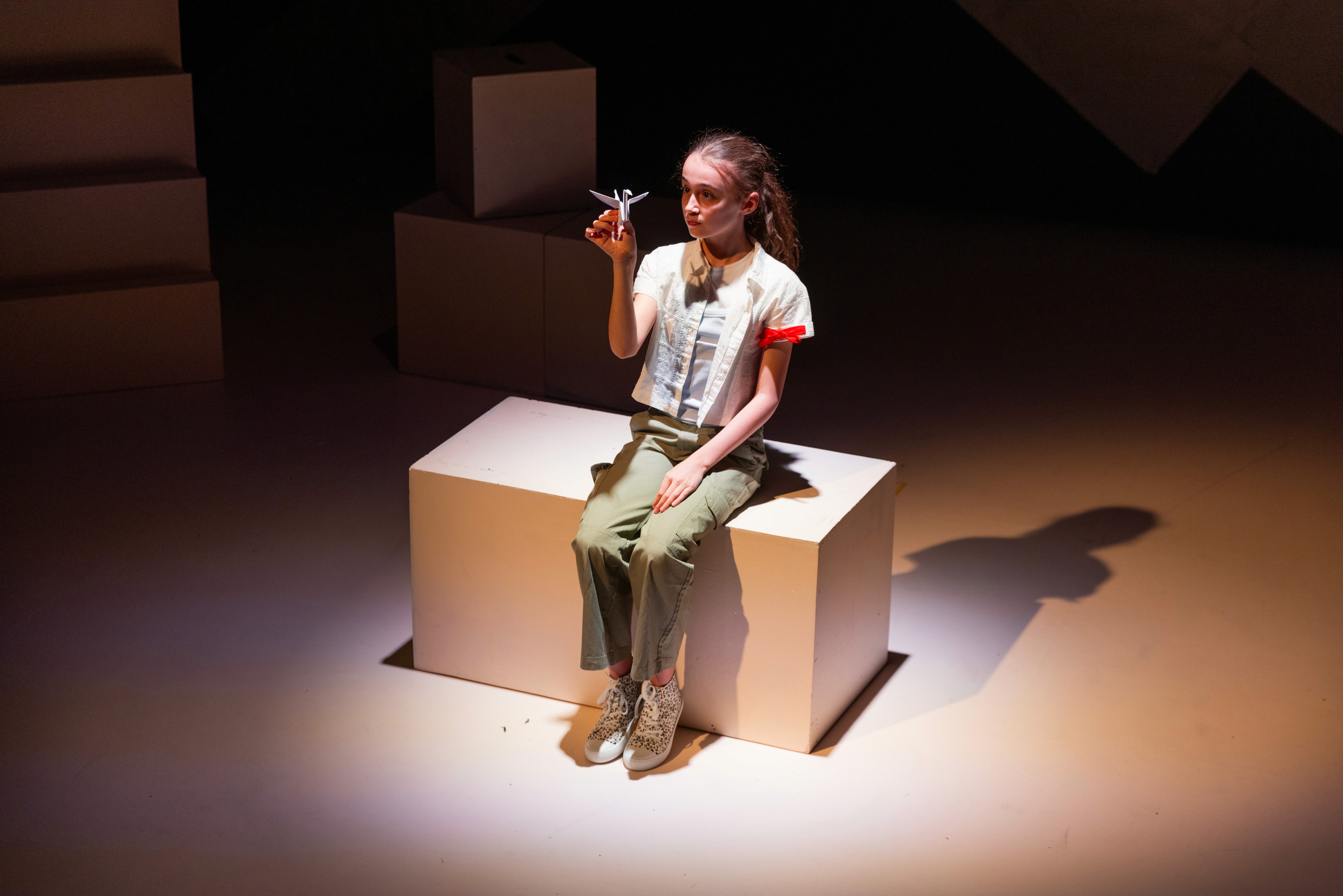 A young performer sits on a white block holding a folded paper crane, lit by a warm spotlight against a minimal stage set with geometric white blocks.